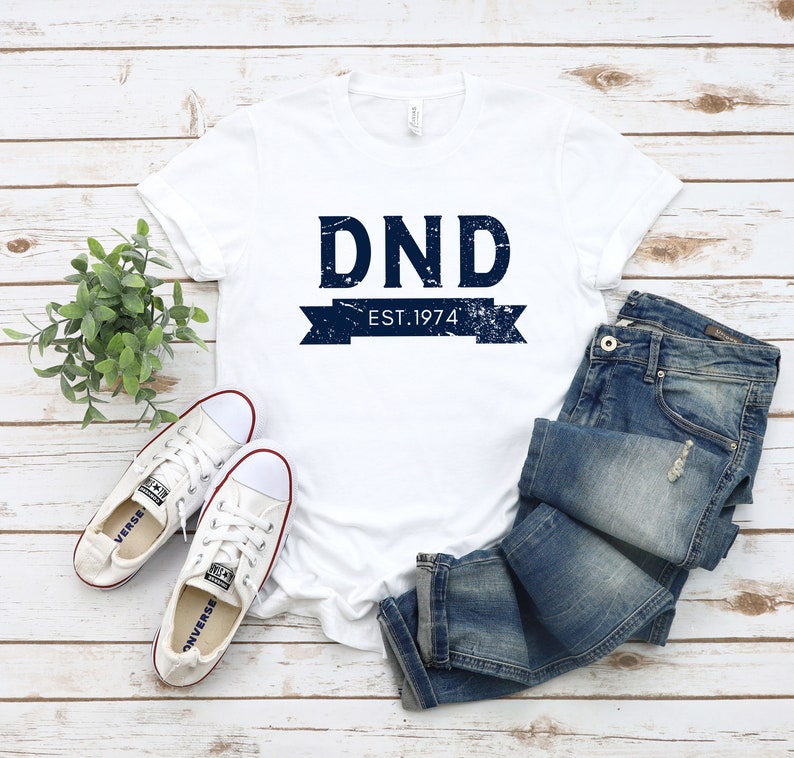 May include: White t-shirt with a blue distressed graphic that says "DND EST. 1974". The shirt is laying on a white wooden surface with a pair of white sneakers and blue jeans.
