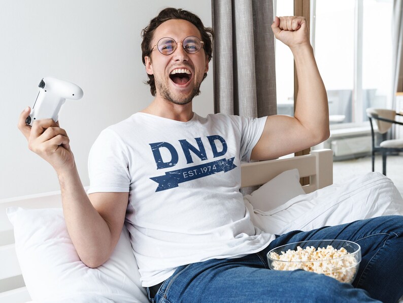 May include: A man wearing a white t-shirt with the text "DND EST. 1974" in blue lettering, sitting on a bed with a white pillow and a bowl of popcorn. He is holding a white video game controller in his left hand and his right arm is raised in the air.