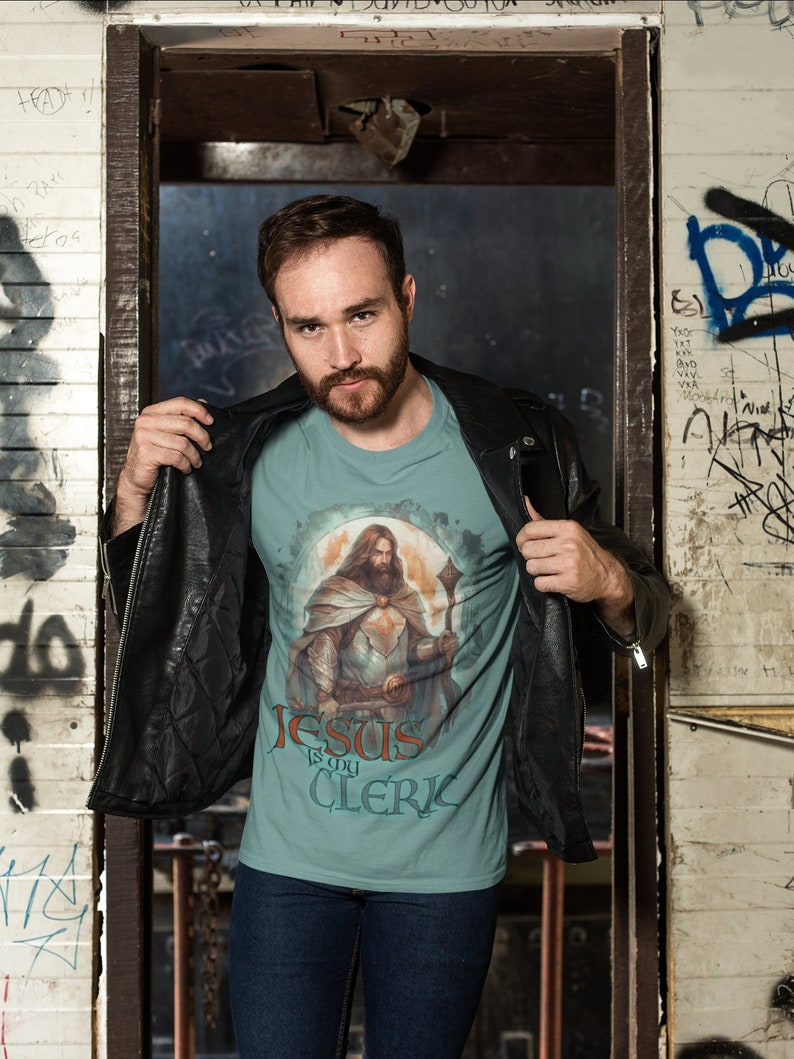 May include: A man wearing a teal blue t-shirt with a graphic of Jesus in armor and the text "Jesus is my Cleric". He is wearing a black leather jacket and blue jeans.