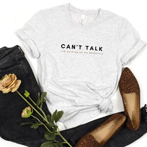 May include: A gray t-shirt with the text "CAN'T TALK I'm working on my backstory" printed in black. The shirt is paired with black jeans and leopard print flats.
