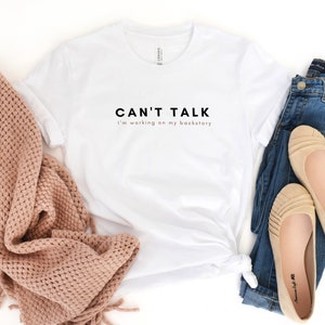 May include: White t-shirt with black text that reads "Can't Talk I'm working on my backstory". The shirt is laid out on a white surface with a pink knitted scarf and blue jeans with brown flats.