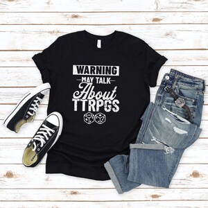 May include: Black t-shirt with white text that reads "WARNING - MAY TALK - ABOUT TTRPGS" and two white dice.