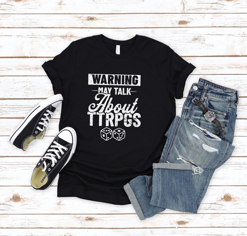 May include: Black t-shirt with white text that reads "WARNING - MAY TALK - ABOUT TTRPGS" and two white dice.