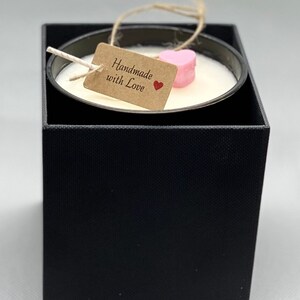 May include: A white candle in a black square box with a pink heart-shaped topper. A brown tag with the text "Handmade with Love" and a red heart is attached to the candle.