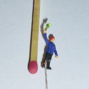 May include: A miniature figurine of a person holding a green object, standing next to a red-tipped wooden matchstick.
