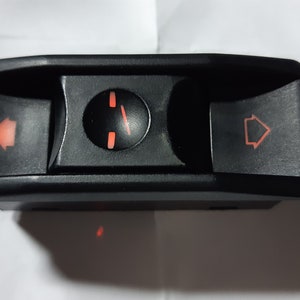 May include: Black automotive switch with three buttons. The left and right buttons have orange arrows, and the centre button has an orange indicator. The switch is likely for hazard lights or indicators.