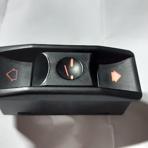 May include: Black automotive control panel with three illuminated orange symbols: a house, a clock, and an arrow. The panel is rectangular with a curved top and bottom, and is likely for a car's interior.