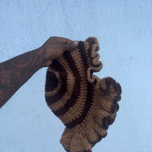 May include: A brown and tan crochet hat with a ruffled brim. The hat features a striped pattern on the crown. The hat is handmade and appears to be soft and comfortable.