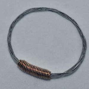 Könnte beinhalten: Ein silbernes Gitarrensaiten-Armband mit goldener Akzent. Das Armband besteht aus mehreren Strängen von Gitarrensaiten, die miteinander verwoben sind.