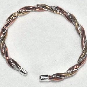 May include: A silver, copper, and gold braided bangle bracelet. The bracelet is twisted and has a smooth, polished finish.