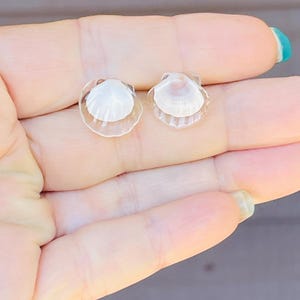 May include: A pair of small, white seashell earrings with clear resin backing. The earrings are shown on a hand.