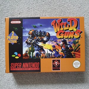 May include: A rectangular Super Nintendo Entertainment System game box for Wild Guns. The orange and black packaging features a graphic illustration of a robotic figure and two characters in a desert landscape, with the text 2 Player Action.