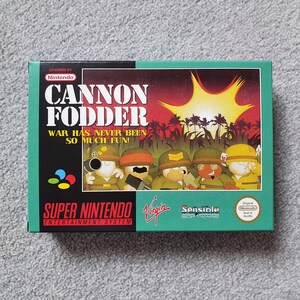 May include: A rectangular Super Nintendo Entertainment System game box for Cannon Fodder. The green box features the title, the text War Has Never Been So Much Fun!, a bright yellow explosion, and five cartoon soldiers in green military uniforms.