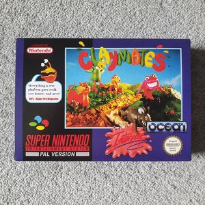 May include: A Super Nintendo Entertainment System game cartridge box for Claymates, a platform game published by Ocean Software. The box is purple with a red splatter design and features a colourful image of the game's characters, which are made of clay.