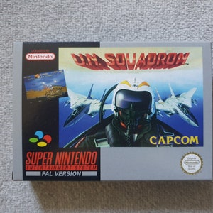May include: A Super Nintendo Entertainment System game cartridge for U.N. Squadron, a classic arcade-style shoot 'em up game. The cartridge is silver with a blue and white label featuring a fighter jet and a pilot in a cockpit. The game is licensed by Nintendo and published by Capcom.