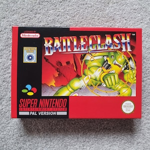 May include: A rectangular box for the Super Nintendo game "Battleclash." The box is red and black, with the game title in white and a green robot illustration. The Super Nintendo logo and "PAL Version" are visible.