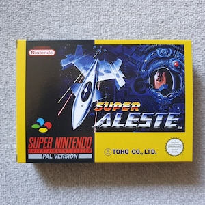 May include: A Super Nintendo Entertainment System video game box for Super Aleste, featuring a yellow and black design with a space fighter ship, a pilot in a helmet, and the text Super Aleste, Super Nintendo Entertainment System, and Toho Co., Ltd.