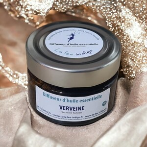May include: A small glass jar with a silver lid, labeled "VERVEINE" and "Diffuseur d'huile essentielle." The jar contains a dark, viscous substance. The background features a shimmering, golden fabric.