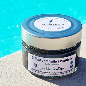 Pode incluir: Um pequeno frasco de vidro com tampa prateada cheio de rocha vulcânica marrom escura. O frasco tem um rótulo que diz "Diffuseur d'huile essentielle Roche volcanique La fée indigo" e inclui o endereço do site da empresa.