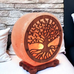 Puede incluir: Una lámpara de sal del Himalaya redonda con un árbol de la vida de madera recortado. La lámpara está encendida y brilla en naranja.