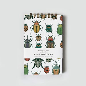May include: A mini notepad with a white background and a repeating pattern of colorful beetles. The notepad has a spiral binding and a paper band that reads "ROOT & BRANCH paper co. MINI NOTEPAD made with love in Knoxville, TN."