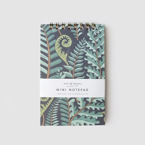 May include: A mini notepad with a fern leaf design. The notepad features a spiral binding and a white band with the text "Root & Branch paper co." and "MINI NOTEPAD". The notepad has a dark background with green and tan fern leaves.