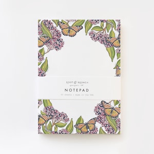 May include: A notepad with a white background and a border of butterflies and flowers. The notepad has a label that says "Root & Branch paper co. NOTE PAD 50 sheets | made in the USA."
