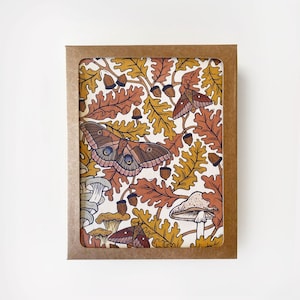 May include: A set of greeting cards in a brown cardboard box. The cards feature a detailed illustration of moths, oak leaves, acorns, and mushrooms in shades of brown, orange, yellow, and cream. The design has a vintage, botanical feel.