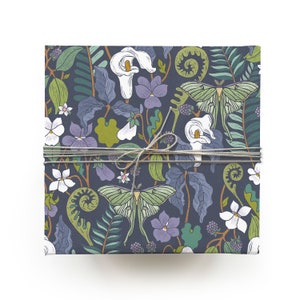 May include: A square gift box wrapped in patterned paper featuring a botanical design. The paper has a dark blue background with illustrations of white calla lilies, green moths, purple violets, and green foliage. The box is tied with twine.