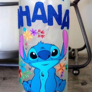 May include: A clear glass bottle with a blue glitter ribbon and the name "DHANA" written in blue paint. The bottle is decorated with a blue Stitch character from Lilo & Stitch, surrounded by colorful flowers.