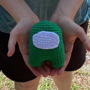 May include: A green crocheted plush toy shaped like the character from the video game Among Us. The toy has a white visor.