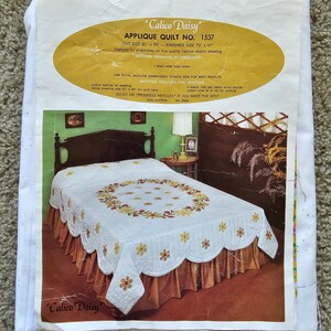 Op de afbeelding: Een vintage appliqué quiltpatroon voor de "Calico Daisy" quilt nr. 1537. Het patroon bevat instructies voor borduren en quilten, en een afbeelding van de afgewerkte quilt op een bed. De quilt is wit met een bloemenontwerp in tinten geel, rood en groen.