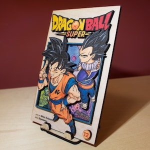 May include: Wooden Dragon Ball Super manga art piece featuring Goku and Vegeta. The artwork is multi-layered with vibrant colors, including orange, blue, and purple. The title "Dragon Ball Super" is prominently displayed. The piece is supported by a wooden stand.