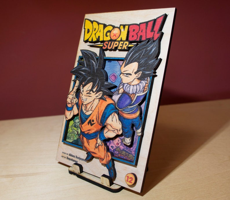 May include: Wooden Dragon Ball Super manga art piece featuring Goku and Vegeta. The artwork is multi-layered with vibrant colors, including orange, blue, and purple. The title "Dragon Ball Super" is prominently displayed. The piece is supported by a wooden stand.