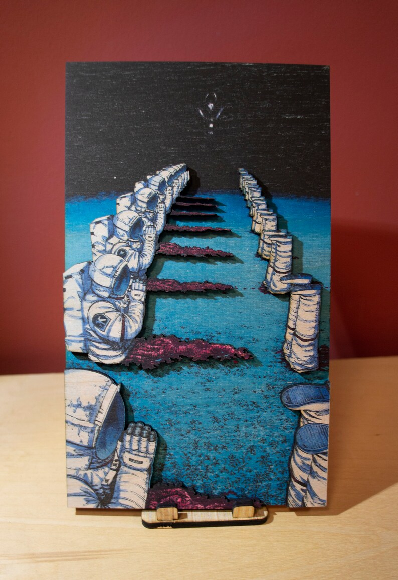 May include: A wooden art piece depicting a line of astronauts in white space suits, walking towards a bright light in the night sky. The artwork features a blue and purple color scheme, with a wooden stand.