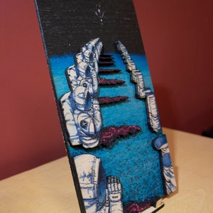 May include: A wooden art piece depicting astronauts in space. The artwork features a dark blue sky with a few stars, a blue and purple landscape, and several astronauts in white and blue space suits. The piece is displayed on a wooden stand.