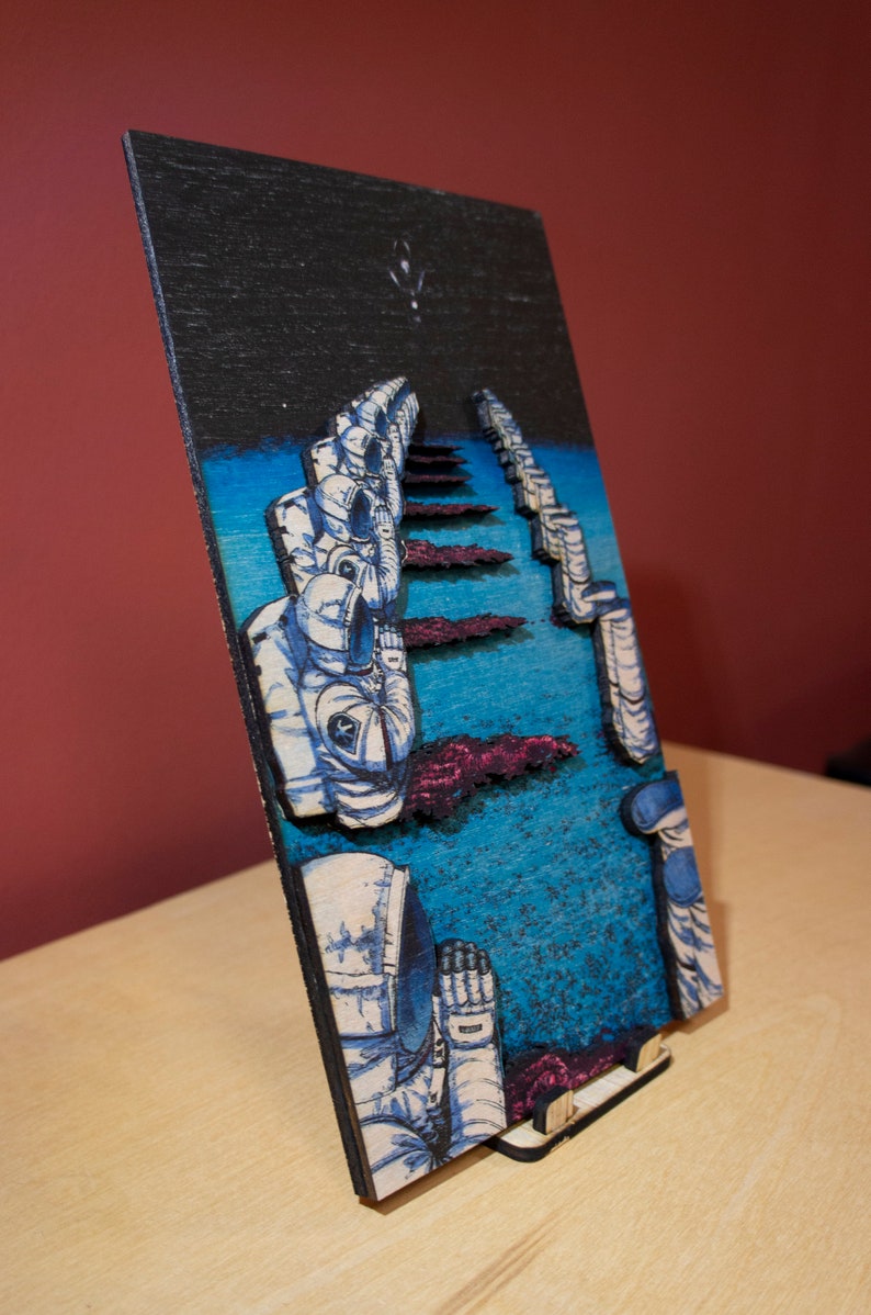May include: A wooden art piece depicting astronauts in space. The artwork features a dark blue sky with a few stars, a blue and purple landscape, and several astronauts in white and blue space suits. The piece is displayed on a wooden stand.