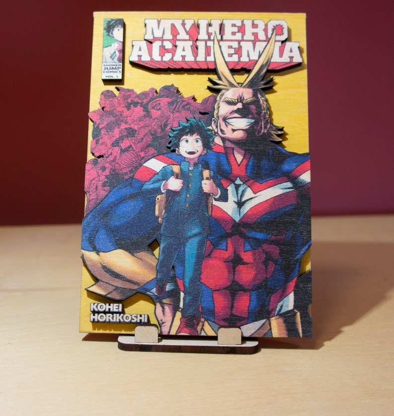 May include: Wooden My Hero Academia manga cover art. The artwork features the title, characters, and author's name. The base is a light brown color, and the artwork is in vibrant colors, including yellow, red, blue, and green.