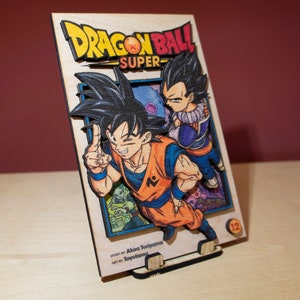 May include: A wooden Dragon Ball Super manga art piece. The artwork features Goku and Vegeta, with the title in yellow and red. The base is made of wood and has a stand. The text "Story by Akira Toriyama, Art by Toyotarou" is visible.