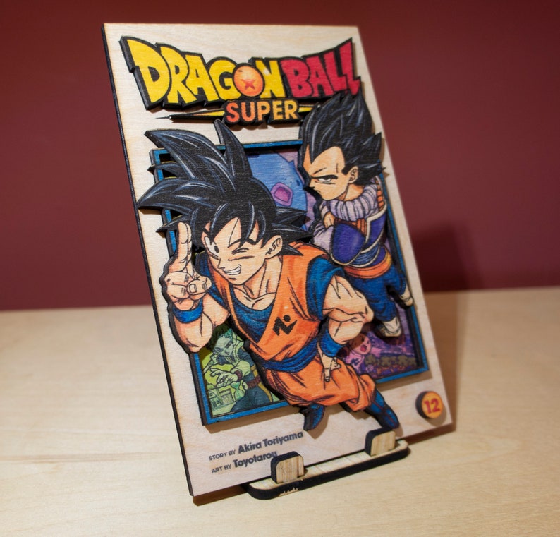May include: A wooden Dragon Ball Super manga art piece. The artwork features Goku and Vegeta, with the title in yellow and red. The base is made of wood and has a stand. The text "Story by Akira Toriyama, Art by Toyotarou" is visible.