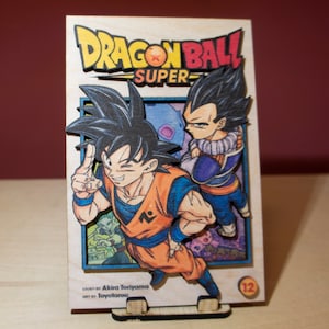 May include: Wooden Dragon Ball Super manga art piece featuring Goku and Vegeta. The artwork is in vibrant colors, with the title and volume number visible. The piece is displayed on a wooden stand.