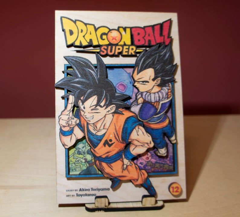 May include: Wooden Dragon Ball Super manga art piece featuring Goku and Vegeta. The artwork is in vibrant colors, with the title and volume number visible. The piece is displayed on a wooden stand.