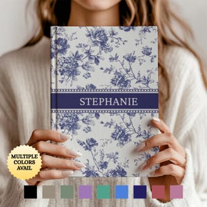 May include: A floral patterned journal with a navy blue band that reads "STEPHANIE". The journal has a cream background with blue floral designs. The image also shows a selection of available colors.