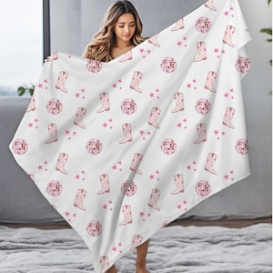 May include: White blanket with a repeating pattern of pink cowboy boots, disco balls, and stars. The blanket is held up, showcasing the design. The boots have a floral design, and the disco balls are pink.