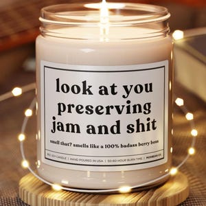 Könnte beinhalten: Eine brennende Sojakerze in einem Glas mit einem weißen Etikett mit der Aufschrift "look at you preserving jam and shit". Die Kerze steht auf einem Holzuntersetzer, umgeben von Lichterketten. Der Text darunter lautet "smells like a 100% badass berry boss."