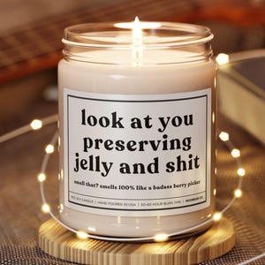 May include: A lit soy candle in a glass jar with a label that reads "look at you preserving jelly and shit." The candle sits on a wooden coaster and is surrounded by string lights. The text below the main text says "smell that? smells 100% like a badass berry picker."