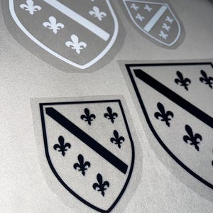 May include: A collection of shield-shaped stickers featuring a coat of arms design. The shields are gray with white and black accents, including fleur-de-lis symbols and diagonal stripes. The stickers vary in size.