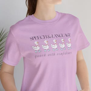 May include: A light purple t-shirt with a graphic of five ducks wearing pink bows and blue boots. The text "SPEECH & LANGUAGE" is above the ducks and "quack with confidence" is below the ducks.