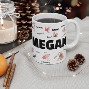 May include: White ceramic mug with a black handle and the name "Megan" printed in bold black letters. The mug is decorated with pink and white floral designs and words describing positive personality traits, such as "kind", "honest", "creative", and "understanding".