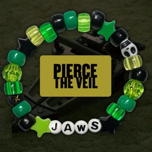 Può includere: Braccialetto di perline verdi, nere e bianche con ciondoli a forma di stella e teschio. Il braccialetto scrive "JAWS" e ha le parole "PIERCE THE VEIL" su uno sfondo dorato.